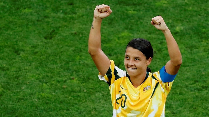 Sam Kerr raises her arms in a strong-woman stance, looking to the crowd with a smile.