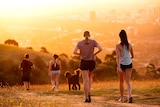 Two groups of people walk a hillside trail with a city in the background at sunset.