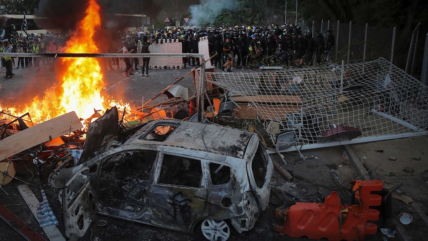 Police in riot gear face a large group as a car and other debris burn in the background