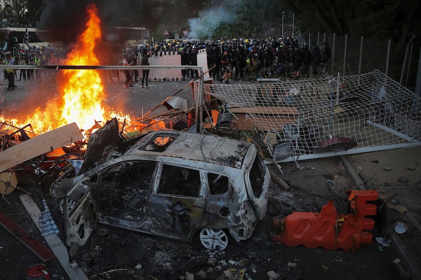 Police in riot gear face a large group as a car and other debris burn in the background