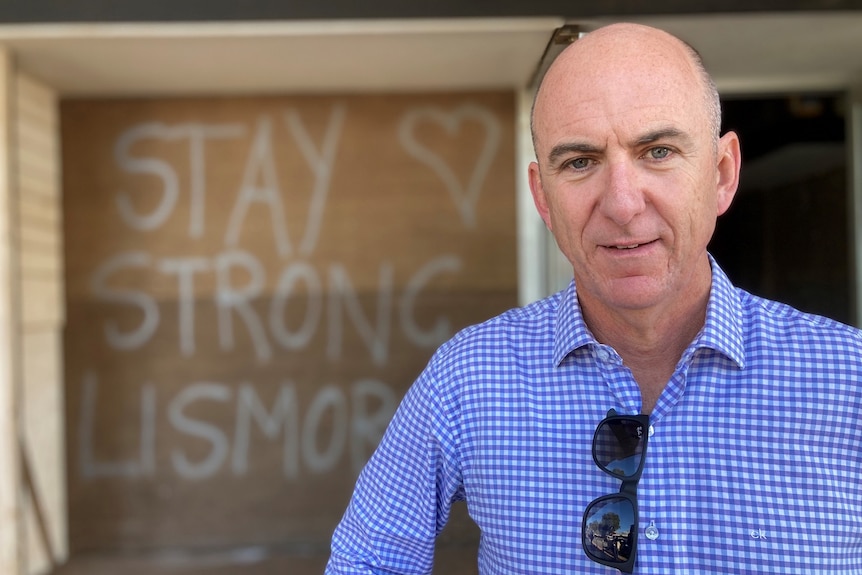 A bald man in a button-up shirt stands in front of a sign that says "stay strong Lismore".