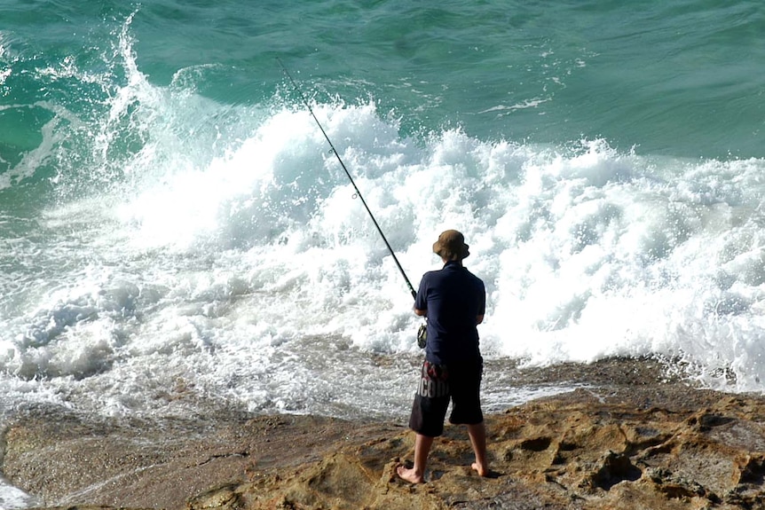 A man in dark shorts, shirt and hat stands alone on rocks fishing in swirling surf