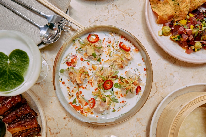 A birds-eye view of a table with a circular plate of kingfish ceviche in coconut milk broth sprinkled with red chilli