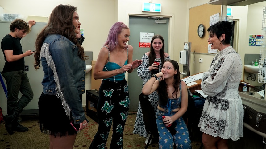 A group of women stand around laughing together in a backstage area 