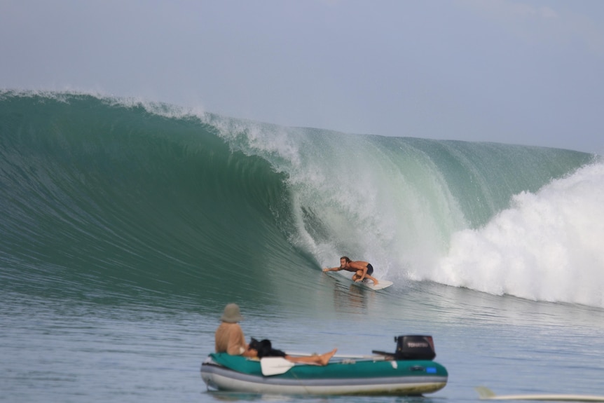 a surfer on a massive wave with his partner in the foreground sitting in a little tender boat