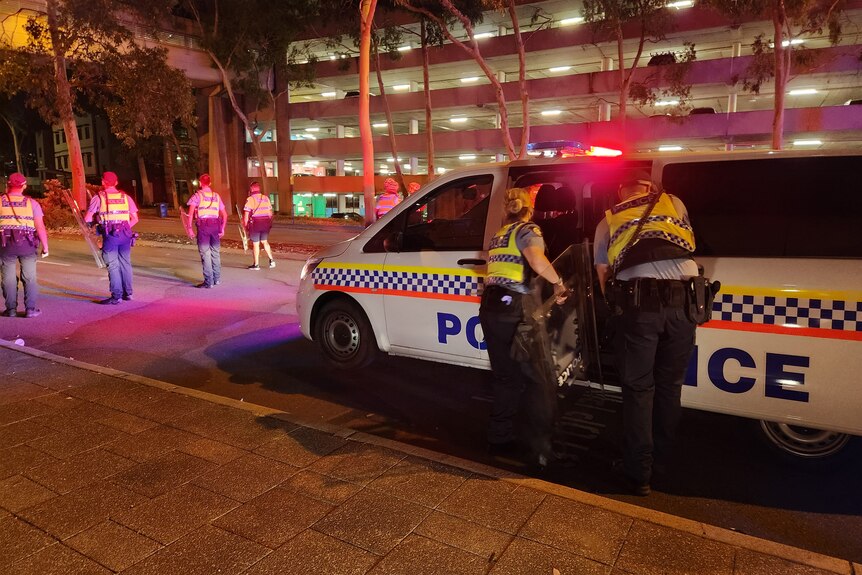 Police officers retrieve riot shields from a police car at night in Perth's CBD with other officers standing across a road.