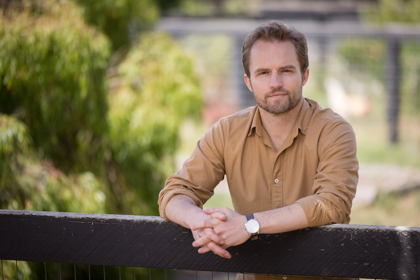 A man wearing a collared shirt leans on a fence.