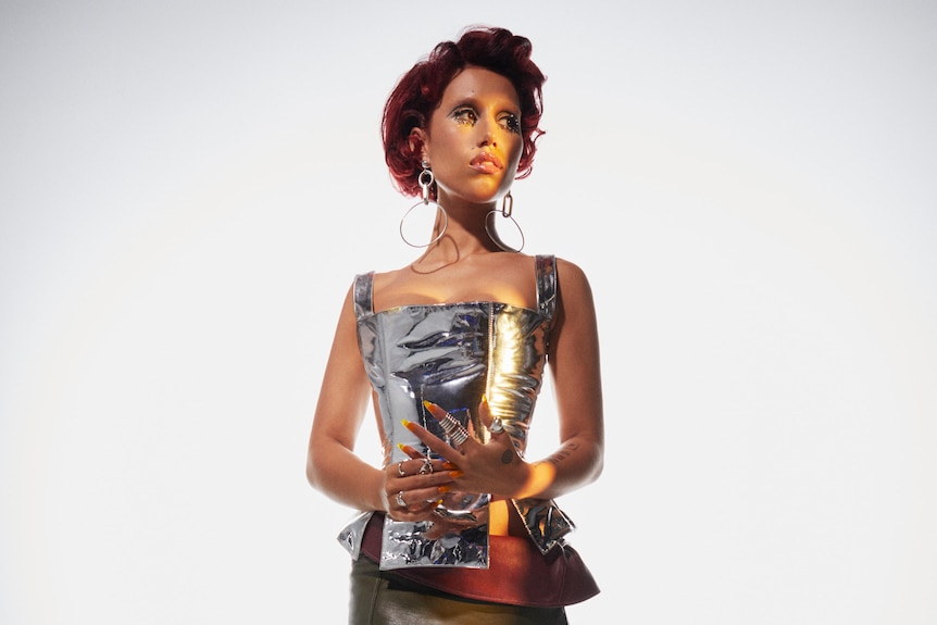 Raye with red hair, long earrings, and silver outfit, looking away from the camera, hands clasped together
