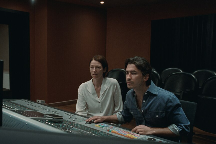 A middle-aged woman with short, combed dark hair sits in a recording studio's control room with a Colombian man