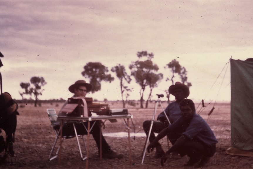 Three men gather around a table set up on an open landscape with a tent and a few small trees behind.