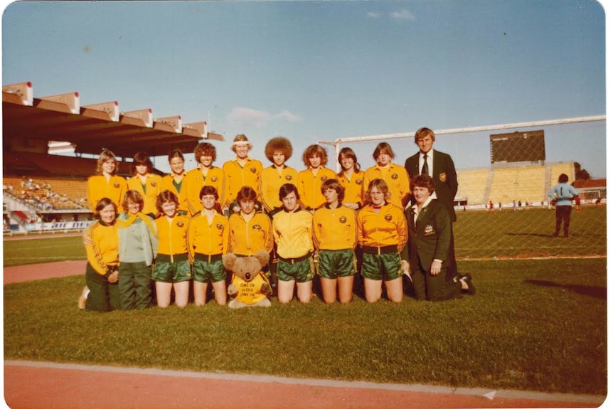 A soccer team wearing yellow jackets and green shorts poses for a photo with a koala toy before a game