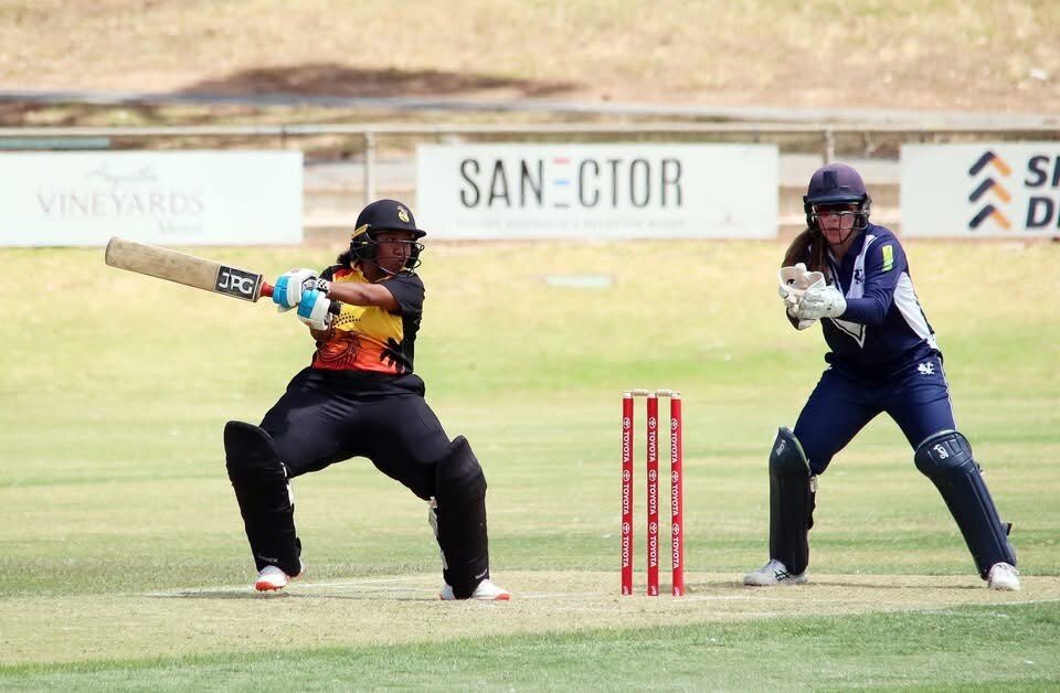 A lady wearing back pants, black helmet and a red, yellow and black top bats on a cricket pitch 