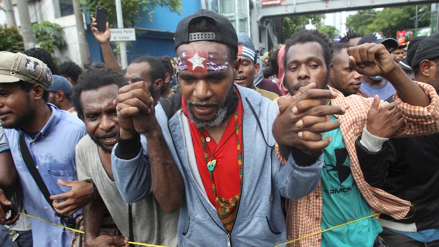 Men at a protest, one with his forehead painted with the West Papuan flag, hold up their hands.