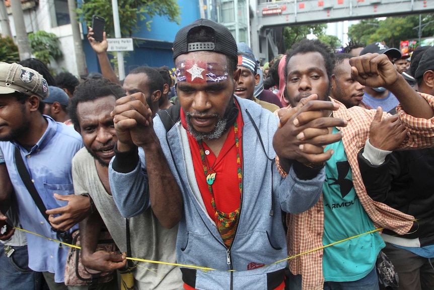 Men at a protest, one with his forehead painted with the West Papuan flag, hold up their hands.