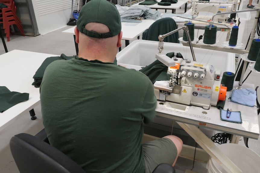 A man wearing a dark green t-shirt sits at a desk with a sewing machine.