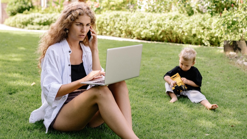 A woman sits on a lawn talking on the phone and looking at a laptop while a toddler plays next to her. 