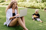 A woman sits on a lawn talking on the phone and looking at a laptop while a toddler plays next to her. 