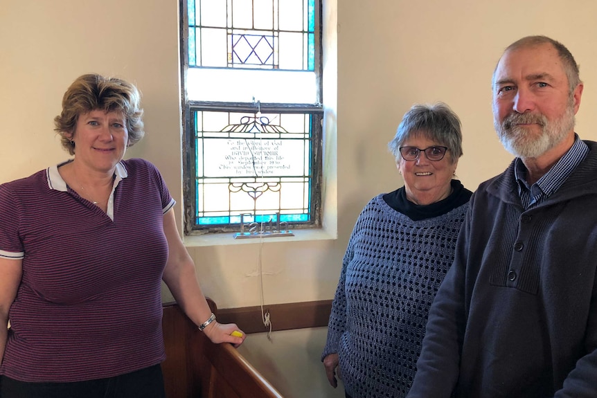 Two women and one man leaning against a church pew, behind a blue stained glass window.