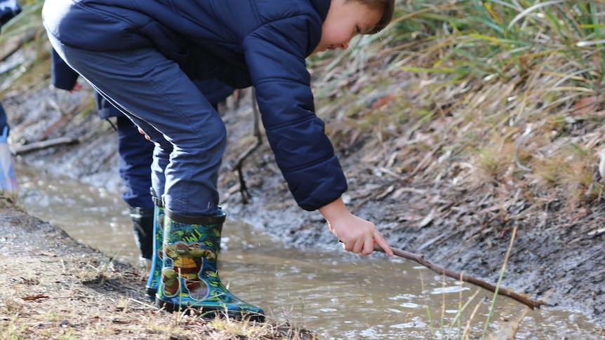 A child in gumboots and jacket pokes a stick at some water