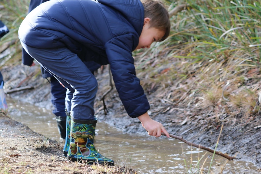 A child in gumboots and jacket pokes a stick at some water