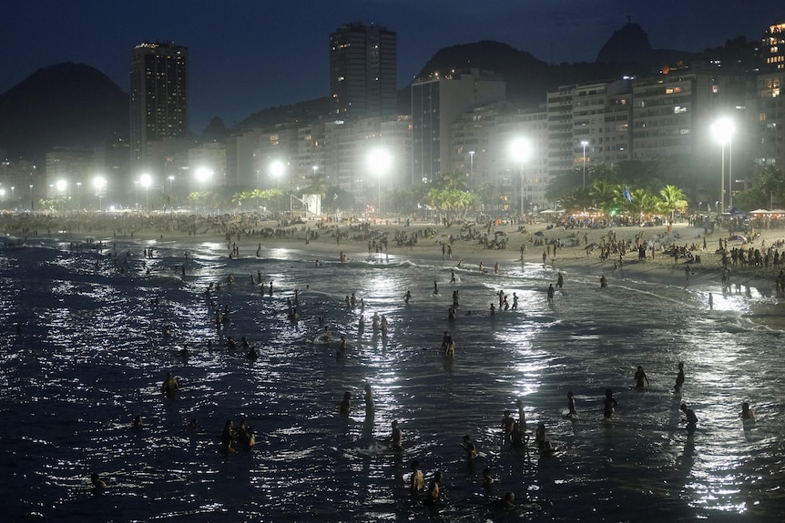 A large crowd of swimmers in the ocean at a city beach at night.