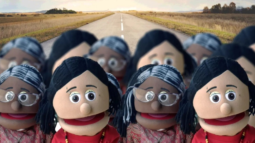 Puppets stand at the front of the picture with horizon and  road behind them.