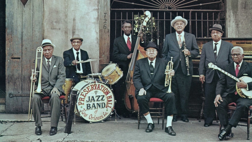 Musicians in suits and fedoras stand with their instruments outside the gate door of an old building.