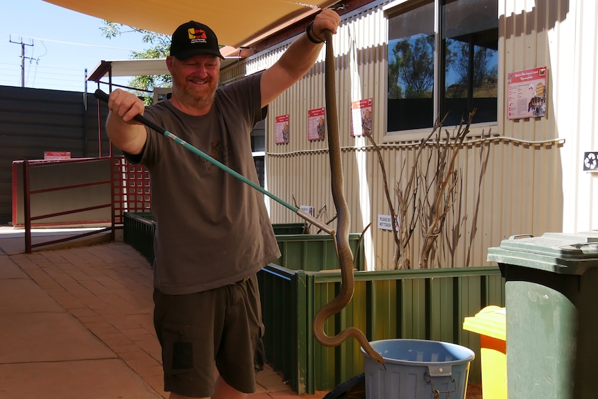 In his left hand a man holds a long brown snake in the air by it's tail, holding a hook in the other hand to support it.