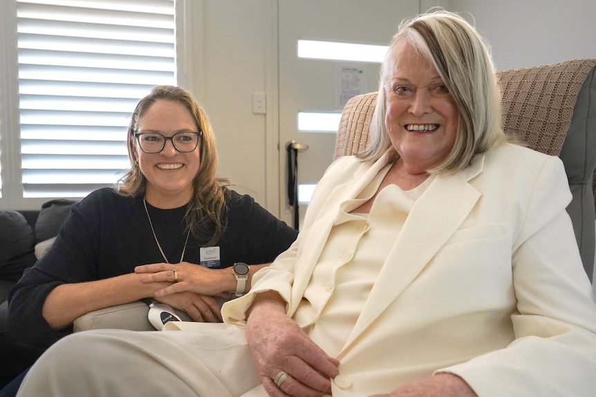 A woman sitting in an armchair, another leans on its arm rest, both smiling