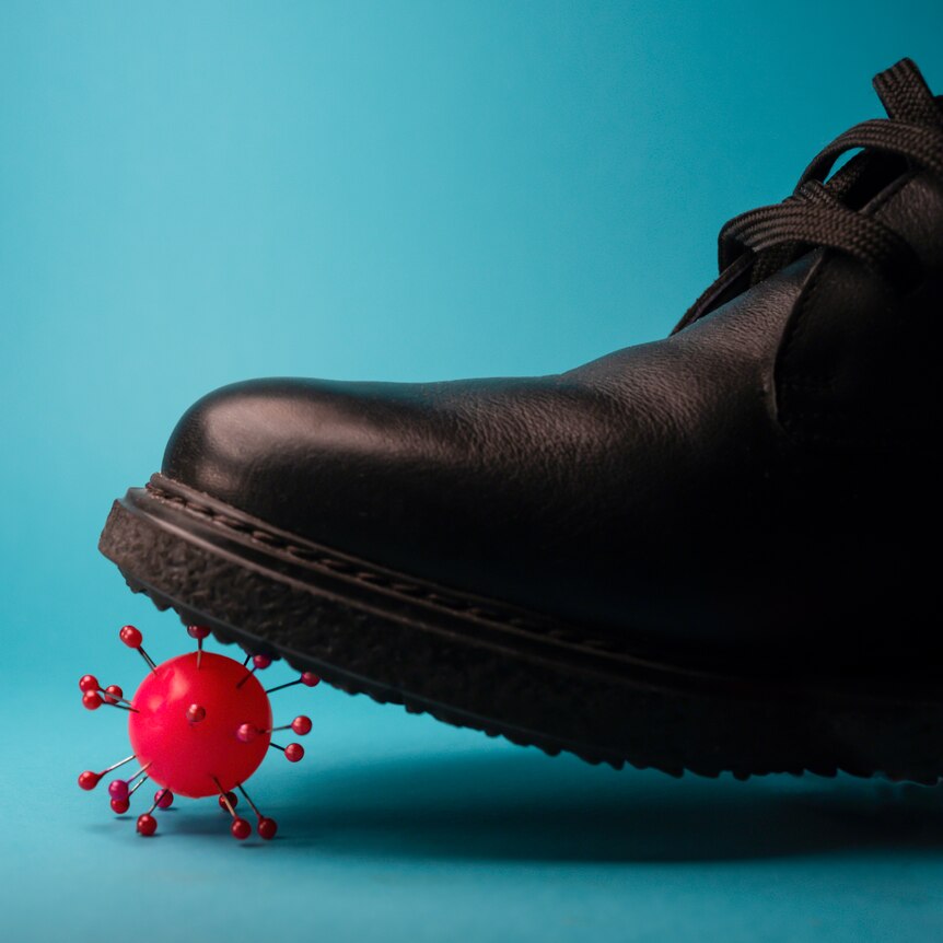 The toe of a black leather shoe steps on the surface of a red ball resembling the molecular structure of a virus.