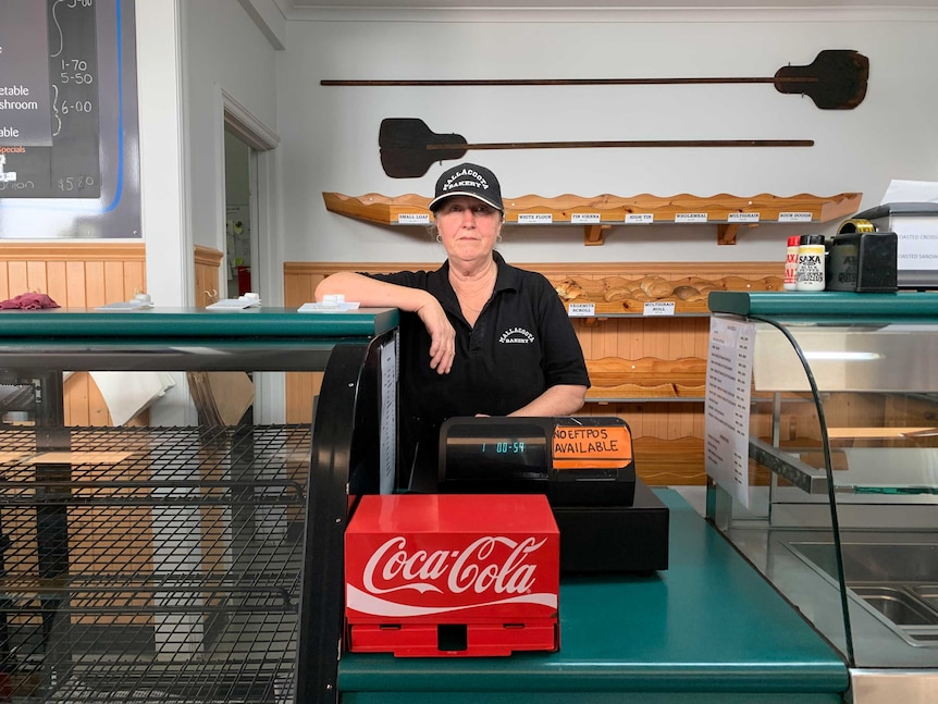 A middle-aged woman standing behind the counter of a bakery. She's wearing a black shirt and black cap.