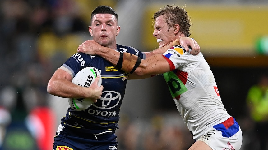A North Queensland NRL player holds the ball as he tries to fend off a Newcastle opponent.