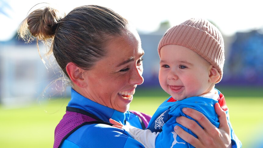A woman athlete wearing blue smiles while holding a baby