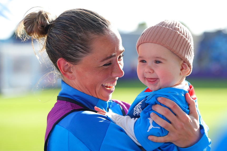A woman athlete wearing blue smiles while holding a baby