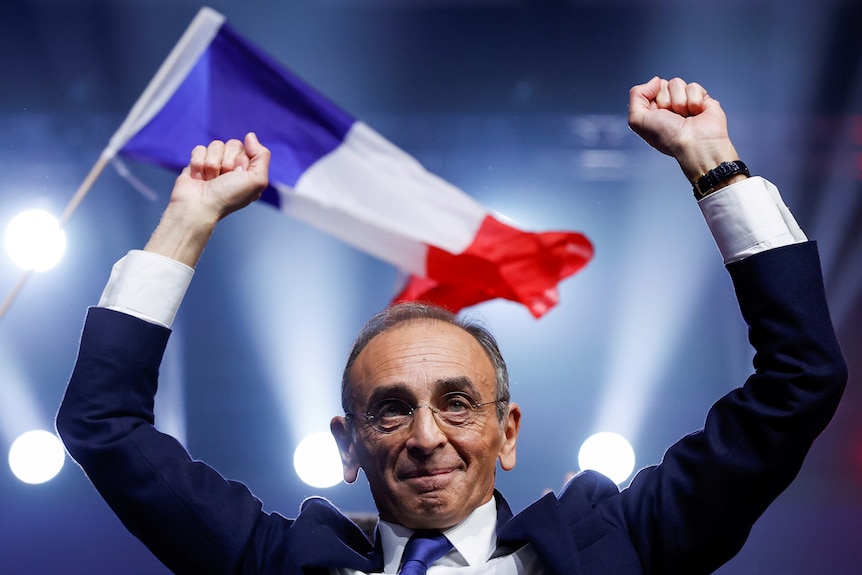 Eric Zemmour with his fists raised in the air with a French flag in the background. 