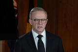 Albanese stands in his office in a black suit and tie with his hands folded.