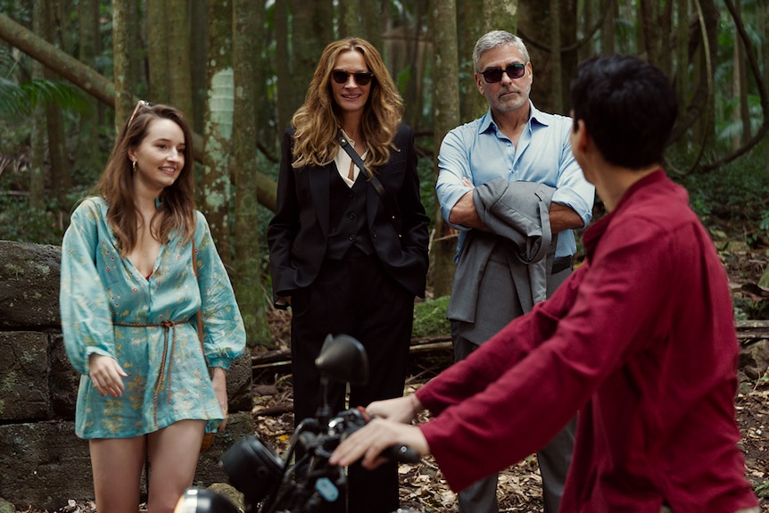 Young woman with brown hair stands in woods with middle-aged red-haired woman and grey-haired man before a young man on a bike.