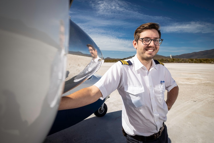 A young man wearing a white shirt with navy and gold epaulettes stands next to the propeller of an aeroplane
