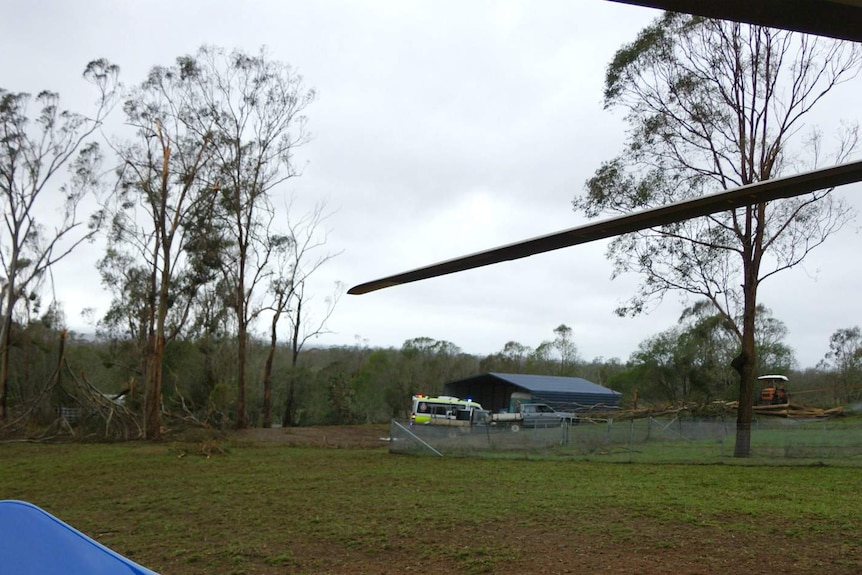 An ambulance in the distance is parked near a shed and an area of fallen trees