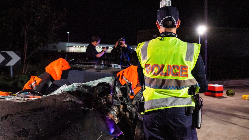 A police officer in a high-vis vest with his back to the camera.  Another officer is in the background taking photos at a crash
