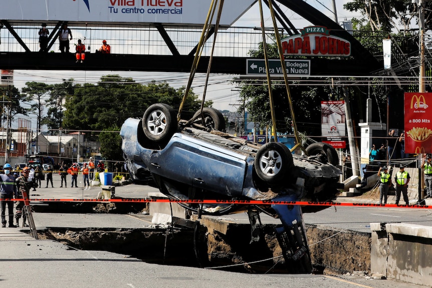A car is suspended by a crane above a sinkhole on a road. Crowds are watching from a foot bridge. 