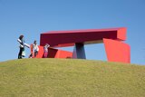 A woman and two children stand in front of a large red, metal sculpture.