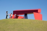 A woman and two children stand in front of a large red, metal sculpture.