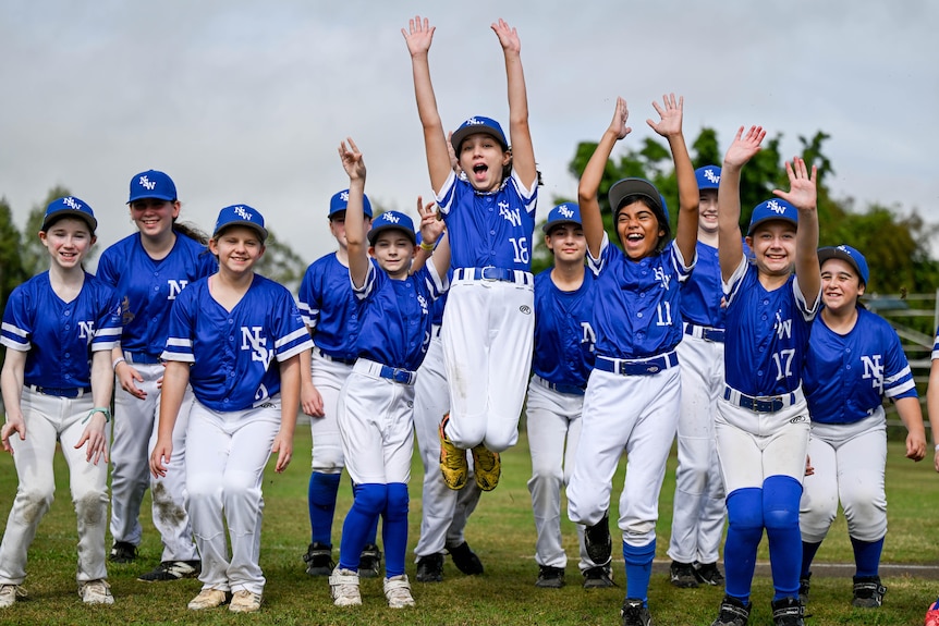 A group of girls in baseball uniforms throw their arms up.