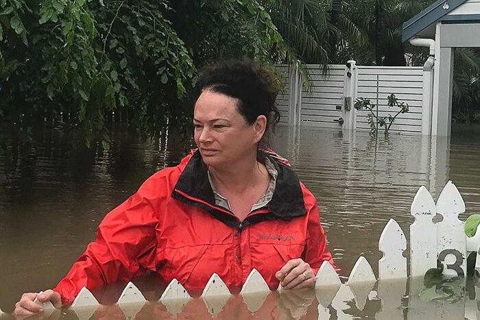 A woman in a red jacket stands in chest high water, the white fence of her garden just peeking out from the surface.