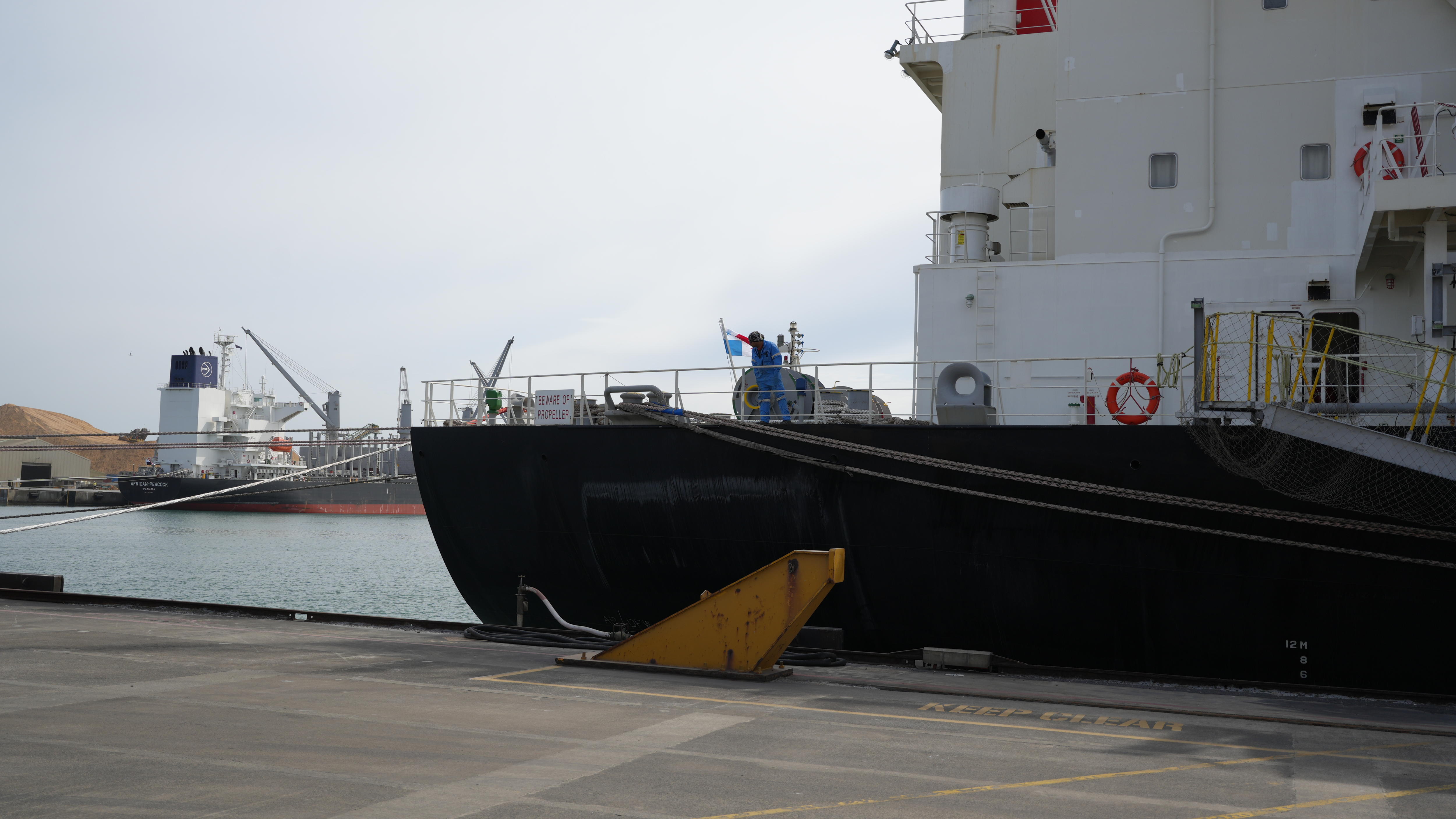 A large black ship is tied to the concrete dock, there are other boats in the distance, a seafarer in a blue uniform is on deck