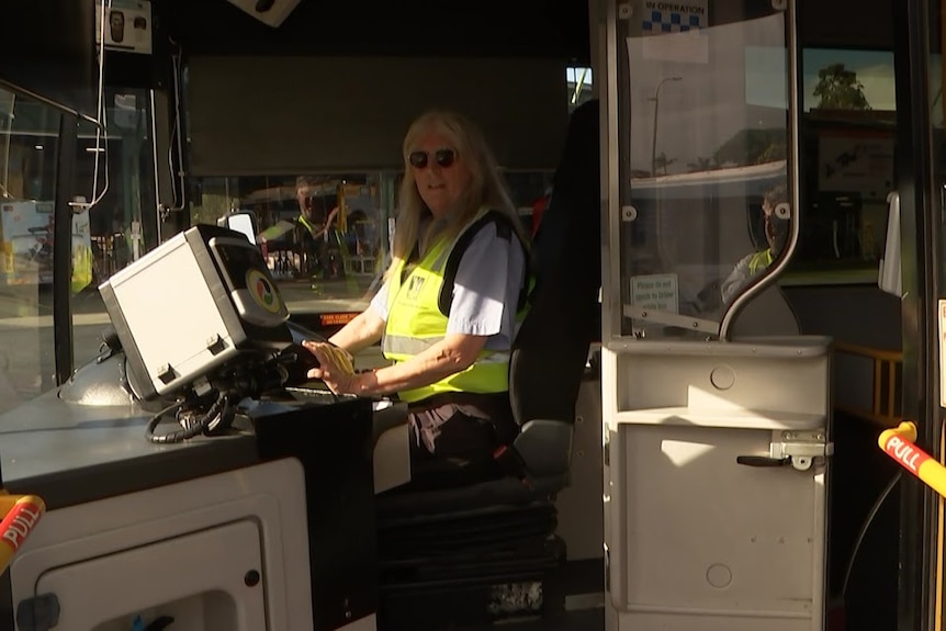 A woman with long blond hair wearing large, dark sunglasses driving a bus. 