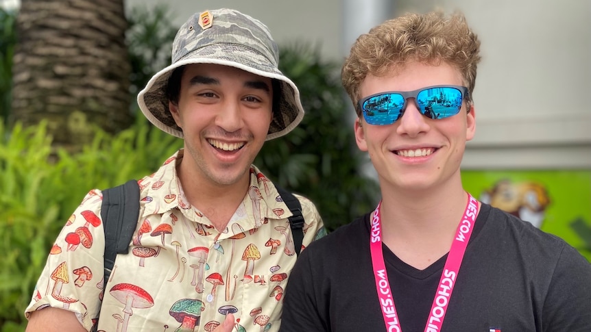 Two teen boys, one wearing a bucket hat and mushroom print shirt, the other with blue sunglasses and pink 'schoolies' lanyard