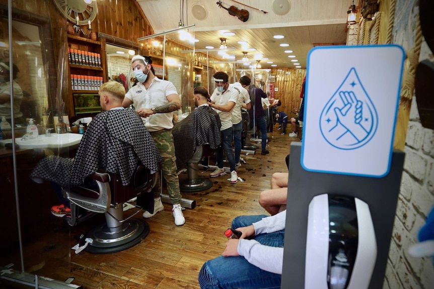 A hand sanitiser dispenser is seen at the door of a barbers shop while men get haircuts inside.