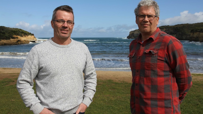 Two men stand in front of a beach.